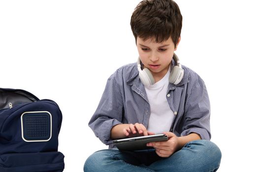 Caucasian smart elementary age student, teen boy in casual denim working on his school project using digital tablet, sitting near his backpack, isolated over white background. Copy advertising space