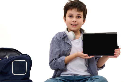 Charming Caucasian teenage boy in casual clothes, showing at camera a digital tablet with empty screen with free space for your ads, sitting near a school bag, isolated on white background. Mockup.