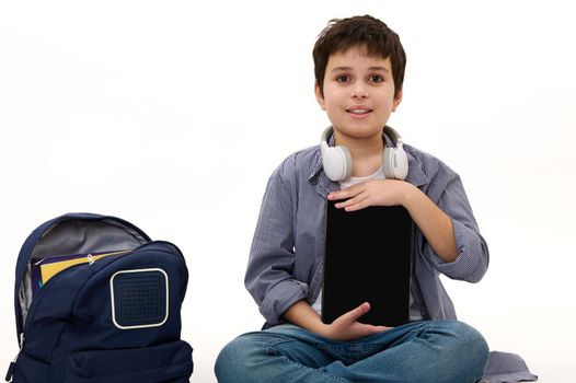 Studio shot of a Caucasian adorable handsome student, teen boy with wireless headphones and backpack, holding a digital tablet with blank screen with copy space for your advertising text. Mockup