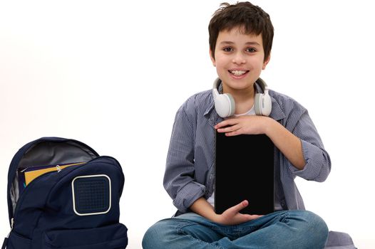 Horizontal shot of a Caucasian adorable handsome student, teen boy with wireless headphones and backpack, holding a digital tablet with blank screen with copy space for your advertising text. Mockup