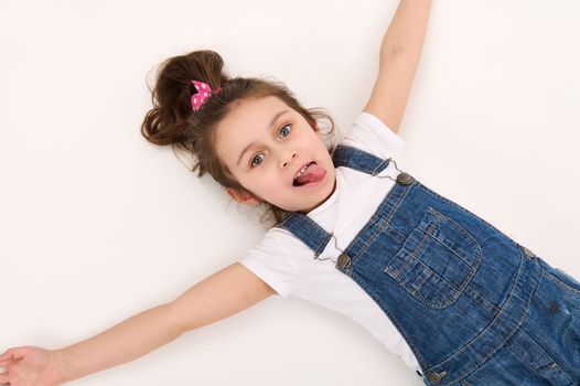 View from above of a Caucasian mischievous child, a lovely baby girl in blue denim overalls and white t-shirt, lying down on a white background. Copy advertising space. Beautiful kids.