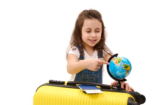 Little girl in denim overalls, with boarding pass and suitcase, points to travel destination on Earth world globe, chooses country to visit, isolated white background. Journey Trip Vacations concept