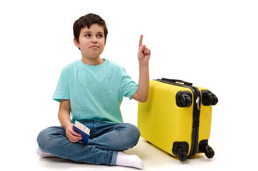 Adorable preteen child, teenage traveler boy in blue denim jeans and casual t-shirt, holding a boarding pass, pointing with index finger at ad space, sitting near yellow suitcase over white background