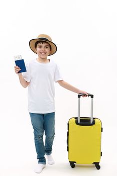 Full size of a teenage boy in casual t-shirt, jeans and straw hat, walking with boarding pass and yellow suitcase on white background. Travel destinations, journey, air trip concepts. Free ad space