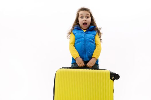 Surprised baby girl with long curly hair in blue jacket, carrying a heavy yellow suitcase, expressing surprise, astonishment, posing with open mouth, looking at camera, isolated on white background.