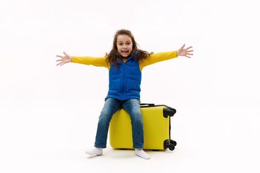 Lovely Caucasian traveler, cute little girl in blue jacket looks at camera, sitting on yellow suitcase with outstretched arms on white background. Happy kid trvelling abroad. Vacations. Travel concept