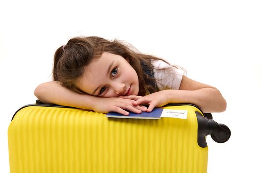 Isolated portrait on white background of lovely baby girl, little traveler with boarding pass and suitcase, looking dreamily at camera, traveling abroad for the weekend. Tourism and air travel concept