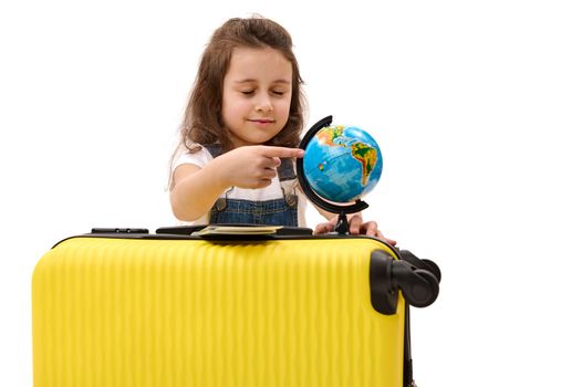 Happy European kid girl 5-6 years old with a suitcase, pointing with her finger at the globe, isolated on white background. Little child passenger traveling abroad for the weekend. Air travel concept