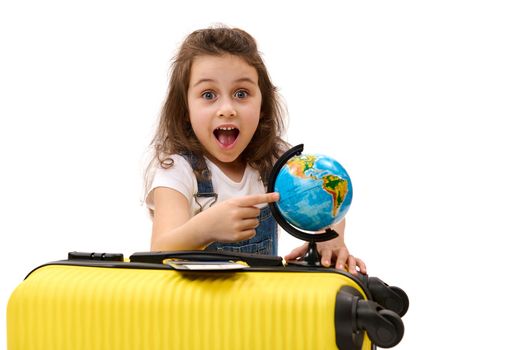 Delightful adorable little girl passenger traveller with yellow suitcase, points to a destination on the globe, surprisely looking at camera, isolated over white background with free advertising space