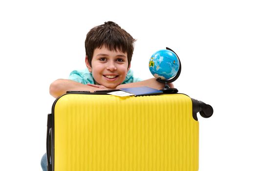 Portrait of a Caucasian happy teenage boy - traveler, passenger with yellow suitcase, boarding pass and globe, smiling a cheerful toothy smile looking at camera, isolated on white background. Ad space