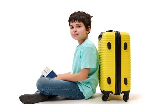 Handsome multi-ethnic teenage boy in casual t-shirt and denim jeans, with boarding pass, leans on yellow suitcase, smiles and looks at camera, isolated on white background. Travel Trip Tourism concept