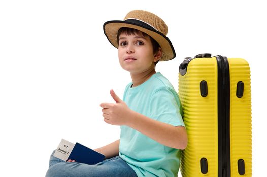 Multi-ethnic handsome teenage boy traveler wearing a turquoise t-shirt, blue jeans and straw hat, posing with yellow suitcase and boarding pass, showing thumb up looking at camera, on white background