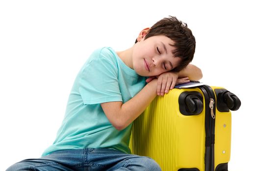 Isolated portrait on white background: adorable teenager traveler boy in blue jeans and casual t-shirt, falling asleep on a yellow suitcase while waiting to board a flight. Air travel Journey concept