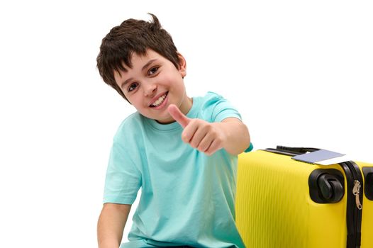 Close-up portrait isolated on white background of a cheerful pre-adolescent passenger boy with yellow suitcase, showing thumb up, cutely smiling to camera. Air travel. Tourism. Journey. Travel concept