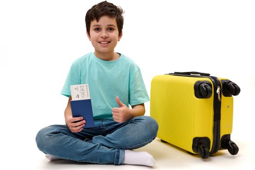 Isolated portrait on white background of teenage boy traveler in casual wear, with boarding pass and suitcase, showing thumb up to camera, traveling abroad for weekend. Tourism and air travel concept