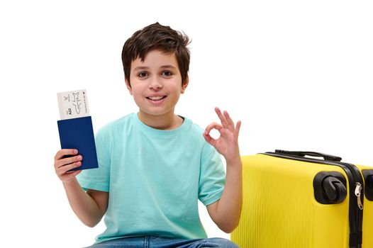 Lovely preteen child, teenage traveler boy in blue denim jeans and casual t-shirt, holding boarding pass, gestures, shows OK sign looking at camera, sitting near yellow suitcase over white background