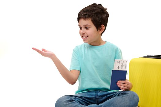 Teenage traveler boy in casual wear with boarding pass, sitting next to yellow suitcase, looking at imaginary advertising space on his hand palm up isolated on white background. Tourism Travel concept