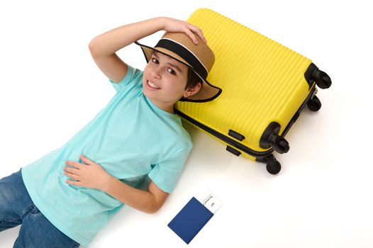 Relaxed preteen child in casual clothes and straw hat, lies on a yellow suitcase, going for summer holidays, isolated portrait over white background. Travel. Tourism. Relaxation. Happy holidays abroad