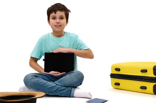 Teenager traveler boy in blue t-shirt, shows at camera a digital tablet with blank screen with space for insert your advertising text or mobile apps, sitting near yellow suitcase on white background