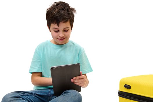 Isolated portrait on white background of a handsome Caucasian teenage traveler boy in casual denim, using digital tablet, browsing websites, checking flight information, sitting near yellow suitcase