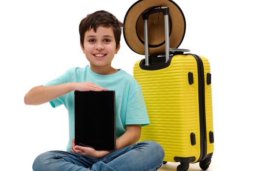 Handsome teenage boy, young tourist traveler, holding digital tablet with empty space - mockup - for advertising text, smiling looking at camera, sitting near his yellow suitcase over white background