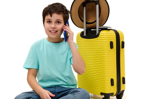 Lovely dark-haired Caucasian teen boy, wearing a turquoise t-hirt and blue casual jeans, looking at camera while talking on mobile phone, sitting over yellow suitcase on white background. Copy space