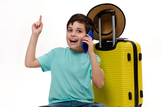 Teenage traveler boy in blue turquoise t-shirt, with yellow suitcase, talking on mobile phone, pointing forefinger at copy space on white background, looking at camera, expressing positive emotions