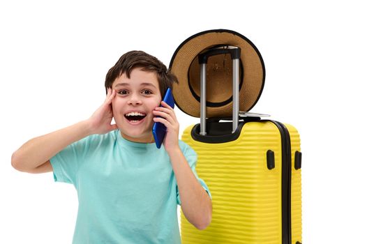 Pre-adolescent child boy, tourist traveler with yellow suitcase, talking on mobile phone, smiling a cheerful toothy smile looking at camera, isolated on white background with free advertising space