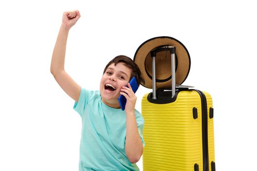 Overjoyed Caucasian teen boy, young traveler tourist in casual wear, raising arms up and clenching fist while having a phone conversation, sitting near yellow suitcase on white background. Copy space