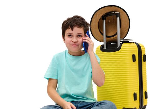 Adorable Caucasian teen traveler boy with yellow suitcase, talking on mobile phone, isolated on white background. Copy advertising . Trip Travel Journey concept