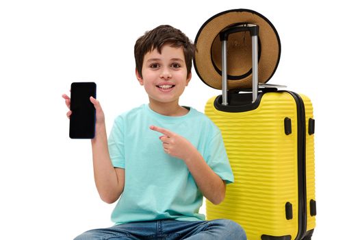 Adorable Caucasian preteen boy in blue casual t-shirt, pointing forefinger at copy space for mobile apps on his modern smartphone, with blank screen, sitting near a yellow suitcase on white background