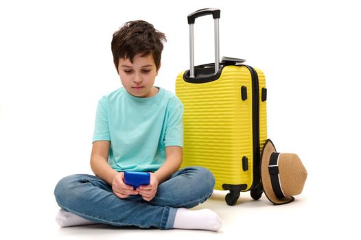 Caucasian teenager boy in blue turquoise casual t-shirt, with yellow suitcase, scrolling news feed, checking social media content, writing sms on his smartphone, isolated on white background. Ad space