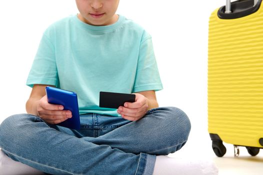 Cropped view of a traveler teenage boy making online payment, using credit card and modern smartphone, sitting near yellow suitcase on white background. Internet banking. Cashless payment concept