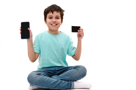 Cheerful Caucasian teenage boy, wearing casual clothes, holding blank smartphone and credit card with copy advertising space, smiling a beautiful toothy smile, looking at camera over white background