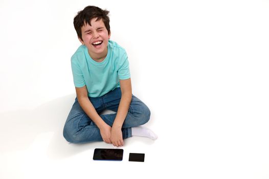 View from above of dissatisfied Caucasian boy, in blue t-shirt and jeans, looking up, expressing sadness and frustration, sitting on white background with modern smartphone and credit card. Copy space