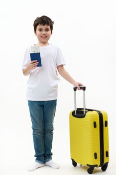 Full size traveler preteen child boy, in casual clothes, holds suitcase and boarding pass, walks isolated on white background. Tourist travel abroad rest getaway Air flight trip journey concept