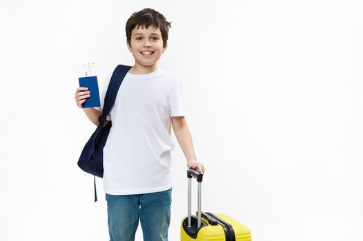 Happy teenage traveler tourist boy in blue jeans and casual t-shirt with backpack, yellow travel bag and boarding pass, smiling a cheerful toothy smile looking at camera, isolated on white background