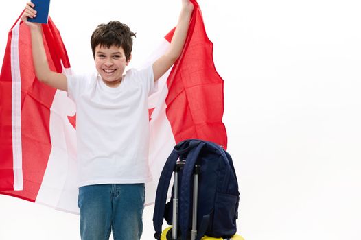 Overjoyed American teenage boy wearing casual t-shirt and blue denim jeans, cheerfully smiling, posing with Canadian flag, boarding pass and luggage isolated over white background. Advertising space