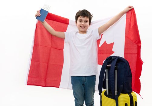 Smart Caucasian teenage boy wearing white t-shirt and blue denim jeans, with backpack, suitcase and boarding pass, carrying Canadian flag, travels to Canada for study exchange experience. Immigration