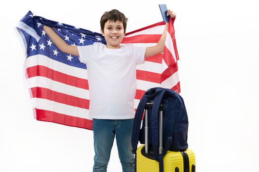 Happy preteen boy in white t-shirt and blue jeans, holds USA flag and boarding pass, smiles at camera, standing next to his backpack on yellow suitcase, isolated over white background, copy ad space