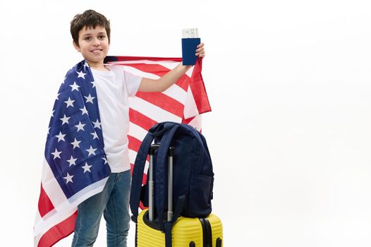 Happy preteen boy in white t-shirt and blue jeans, holds USA flag and boarding pass, smiles at camera, standing next to his backpack on yellow suitcase, isolated over white background, copy ad space