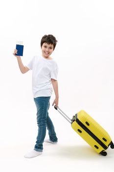 Full length portrait of a handsome teenage boy in blue denim and white t-shirt, with boardig pass and suitcase going on summer vacation, isolated on white background. Holiday, travel, kids concept