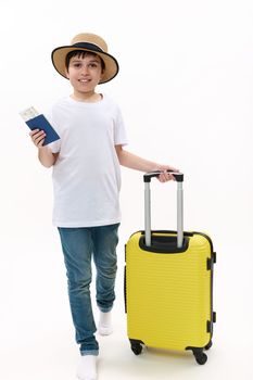 Full size of traveler child, teenage boy wears casual clothes, holds suitcase and boarding pass, walks isolated on white background. Tourist travel abroad rest getaway. Air flight trip journey concept