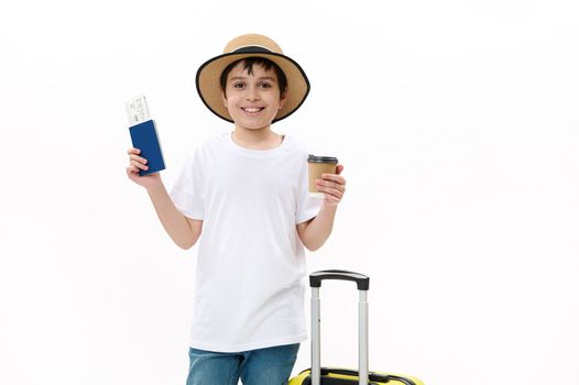 Happy teenage traveler tourist boy in blue jeans, casual t-shirt and straw hat with travel bag, holds a takeaway hot drink in disposable cardboard cup and boarding pass, isolated over white background