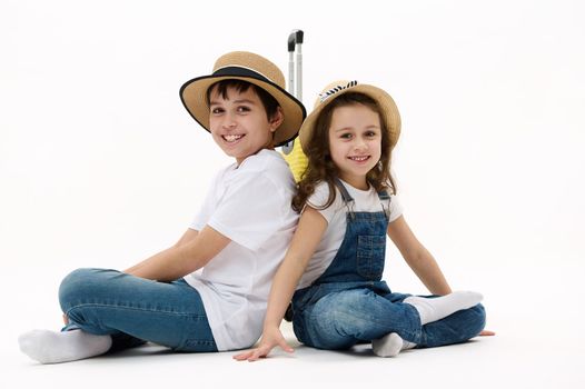 Adorable diverse children, preteen boy and little girl sit with their backs to each other against yellow suitcase, cutely smile on camera, isolated on white background. Kids traveling. Family journey