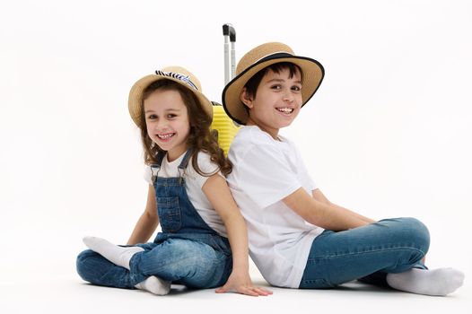 Lovely kids, brother and sister sit with their backs to each other in lotus pose, with yellow suitcase, cutely smile on camera, isolated on white background. Children travel abroad for weekend getaway