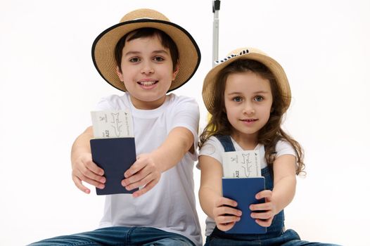 Happy journey. Family of travelers brother and sister wearing casual denim, white t-shirt and straw hat, with suitcase, showing on camera air tickets and passports, isolated on white background