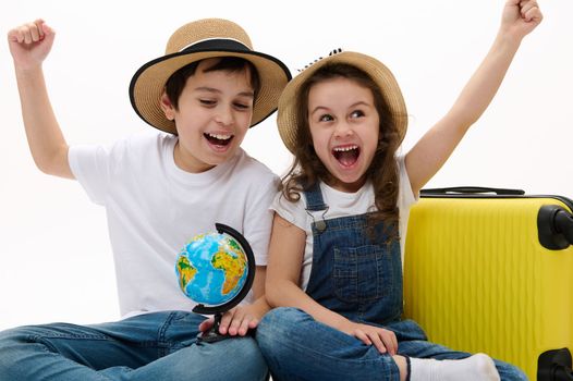 Overjoyed kids, little girl and preteen boy in casual wear, young travelers experience happiness while going for summer vacations, sitting with globe and yellow suitcase, isolated on white background