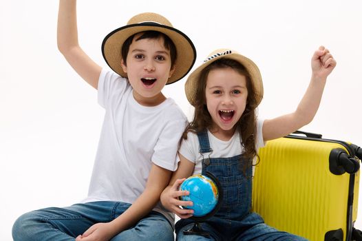 Overjoyed kids clenching fists from happiness, enjoying the upcoming travel abroad, sitting against a yellow suitcase with Earth map globe, on white background and cheerfully smiling looking at camera