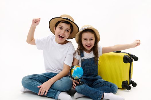 Young tourists boy and girl going abroad for weekend getaway, choosing destination on the globe map, clenching fists from excitement and happiness, sitting near yellow suitcase on white background